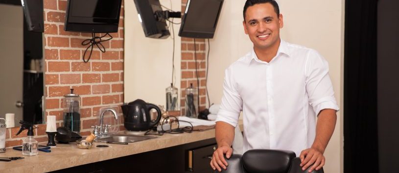 Handsome young barber standing behind a barber chair and greeting clients with a smile