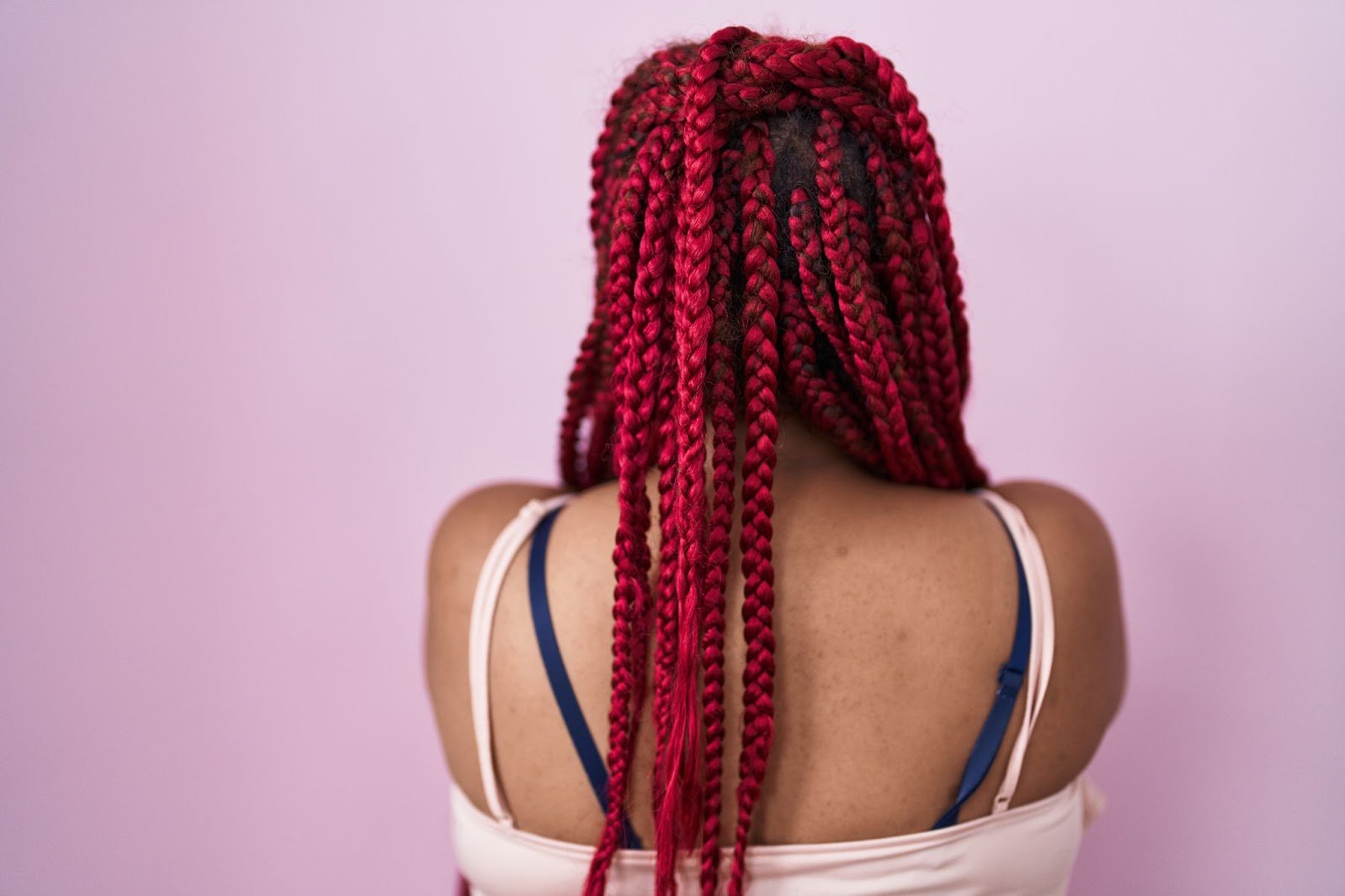 How To Braid African Hair: A Guide for New Cosmetologists 5 Back view of a woman with long, red box braids against a light pink background.