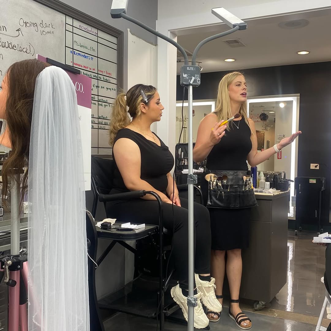 Makeup instructor explaining techniques to a student during a live demonstration at a beauty school, with a mannequin in a bridal veil nearby.