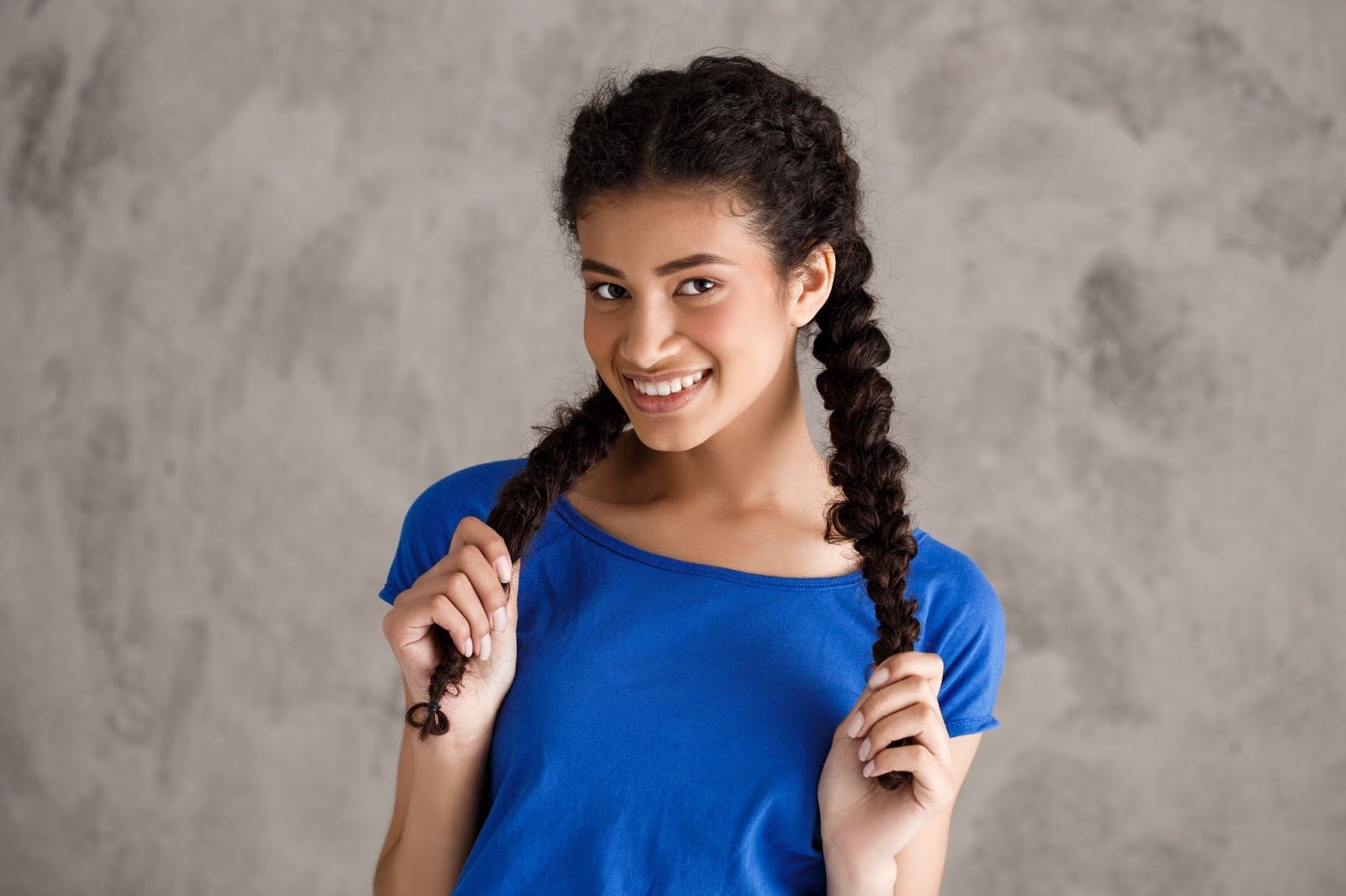 How To Braid African Hair: A Guide for New Cosmetologists 7 Smiling woman holding two French braids, wearing a blue shirt in front of a textured background.