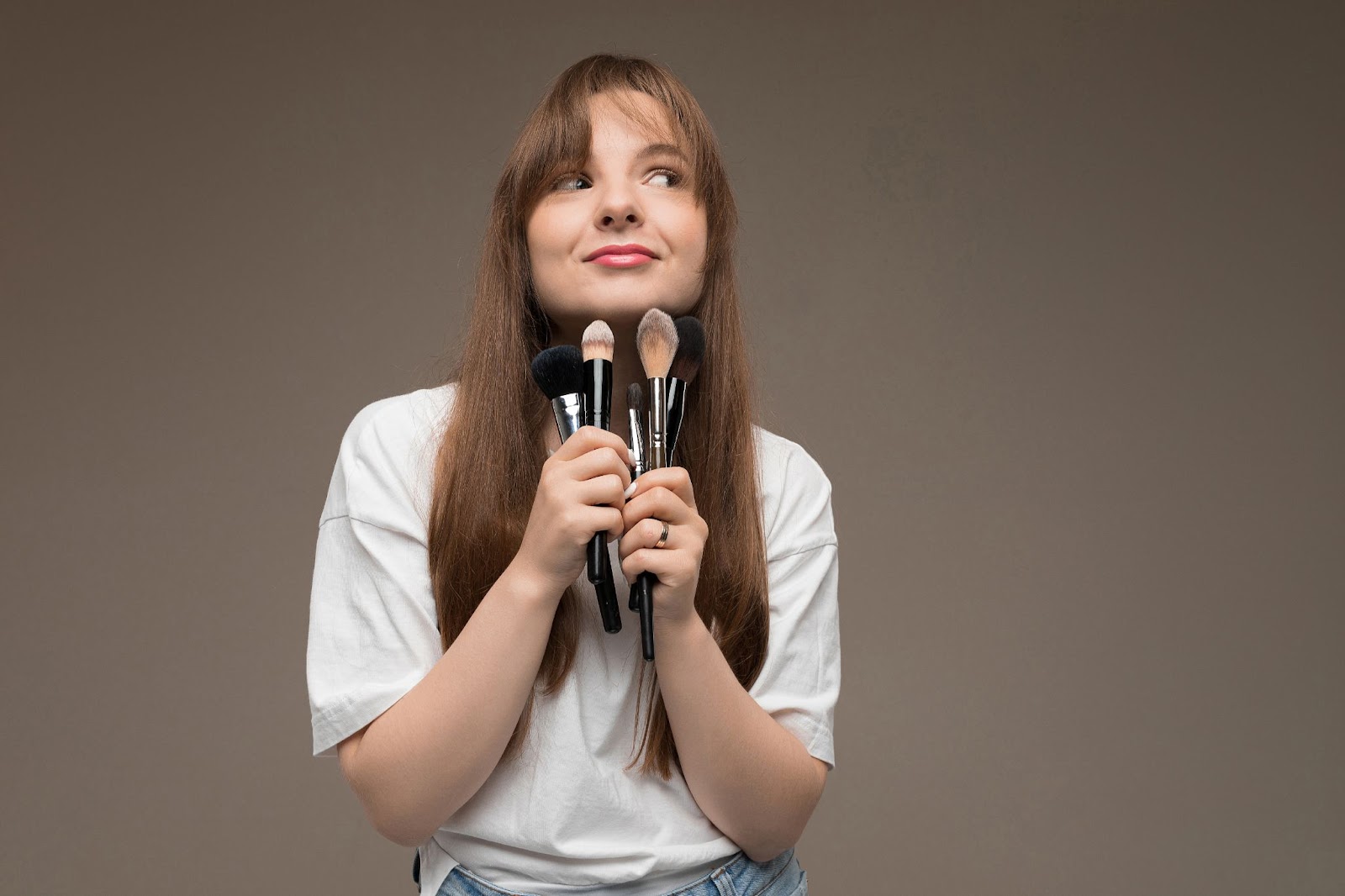 Girl holding beauty and cosmetology brushes in her hand and looking at other direction while smiling softly. 
