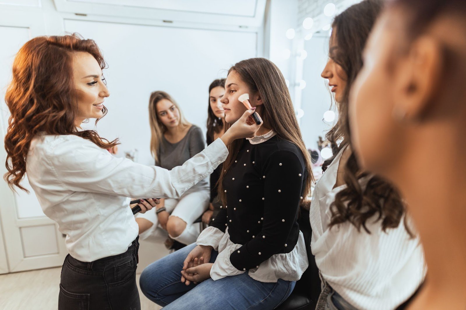 Beauty instructor applying blush to a student model during a makeup class, with other students watching in a modern studio.