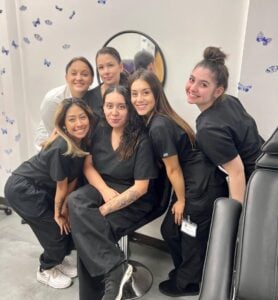 Group of smiling beauty school students in black uniforms posing together in a classroom with butterfly decorations.