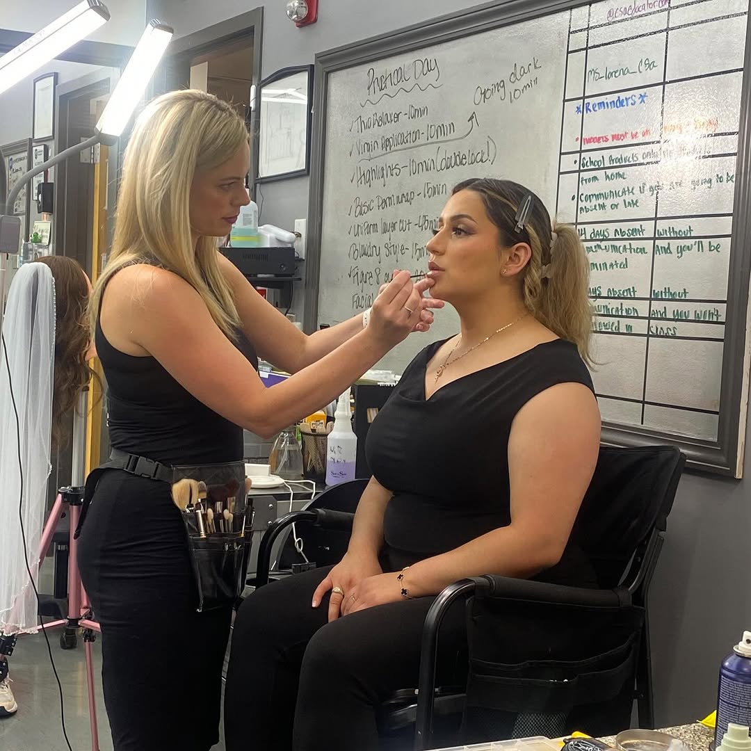 How to Choose a Beauty School: Making The Right Decision 1 Makeup student applying lipstick to a woman sitting in a salon chair, with a makeup brush belt and whiteboard instructions in the background.
