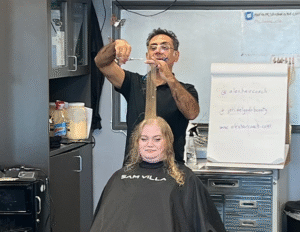 Instructor demonstrating haircutting techniques on a student in a beauty school classroom, with a flip chart and whiteboard in the background.