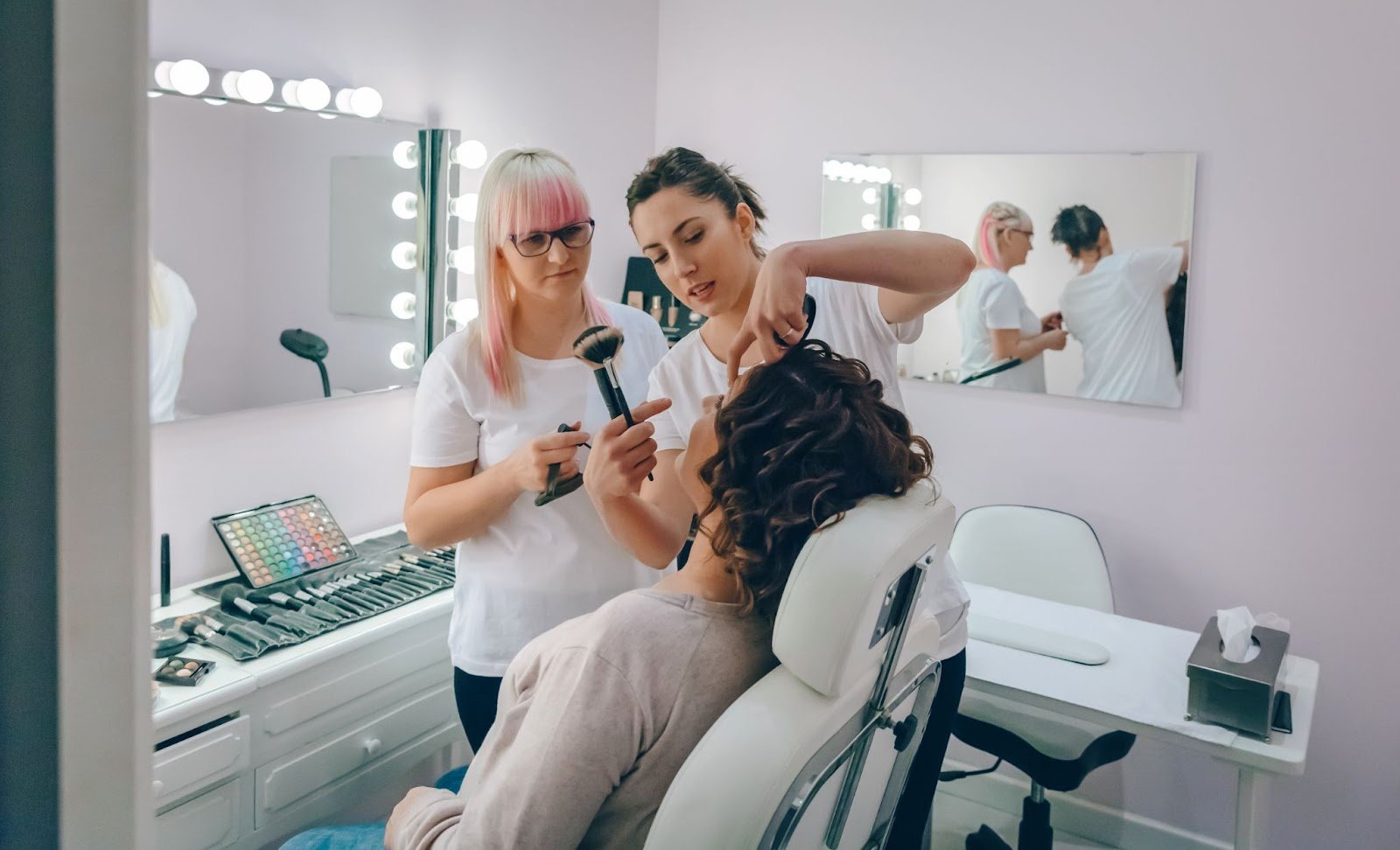 Two makeup artists working together on a client in a professional beauty studio with bright vanity lighting.