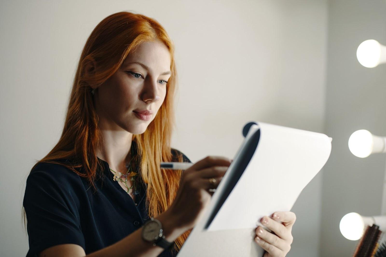 Cosmetology State Board Exam: How to Prepare 6 Red-haired woman reviewing notes on a clipboard in a cosmetology classroom or studio