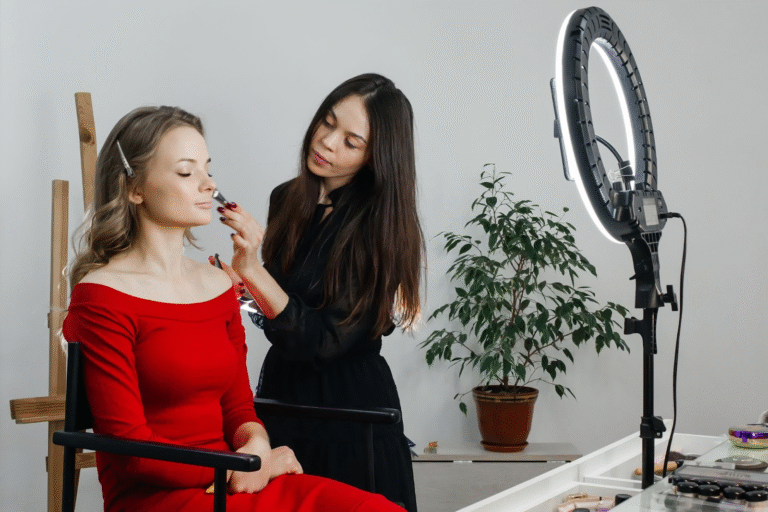 Makeup artist applying foundation to a client in a red dress under a ring light setup.